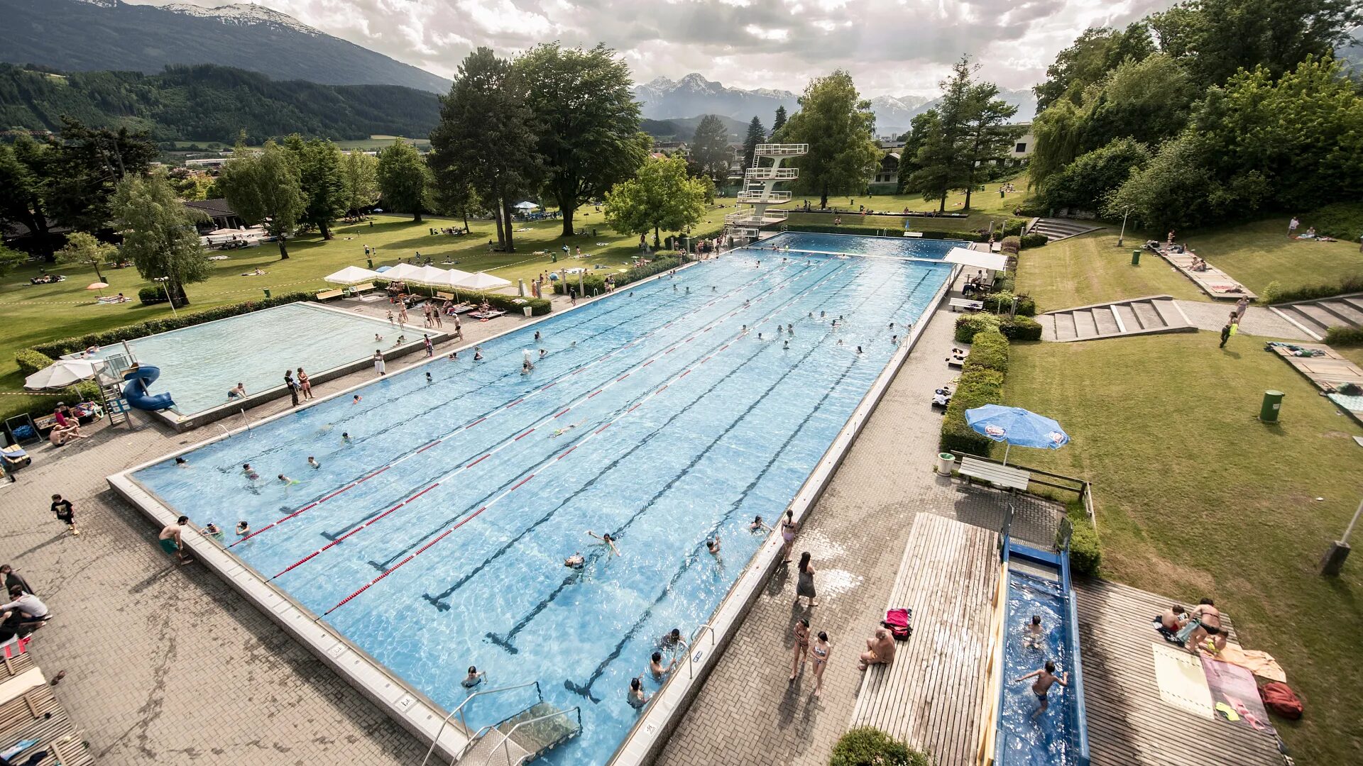 Freibad mit großem Schwimmbecken, Sprungturm, Planschbecken und Liegewiese vor bewaldeten Bergen