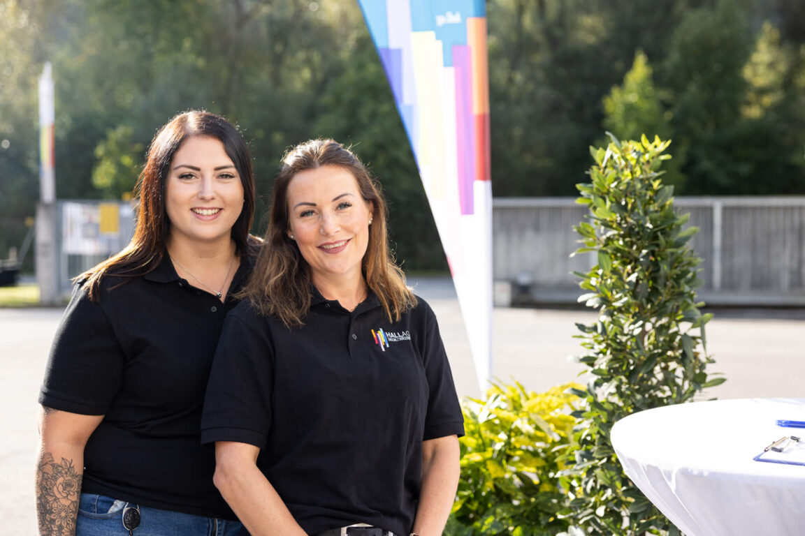 Zwei Mitarbeiterinnen der HAll AG stehen nebeneinander im Freien, beide tragen schwarze Poloshirts mit Firmenlogo.