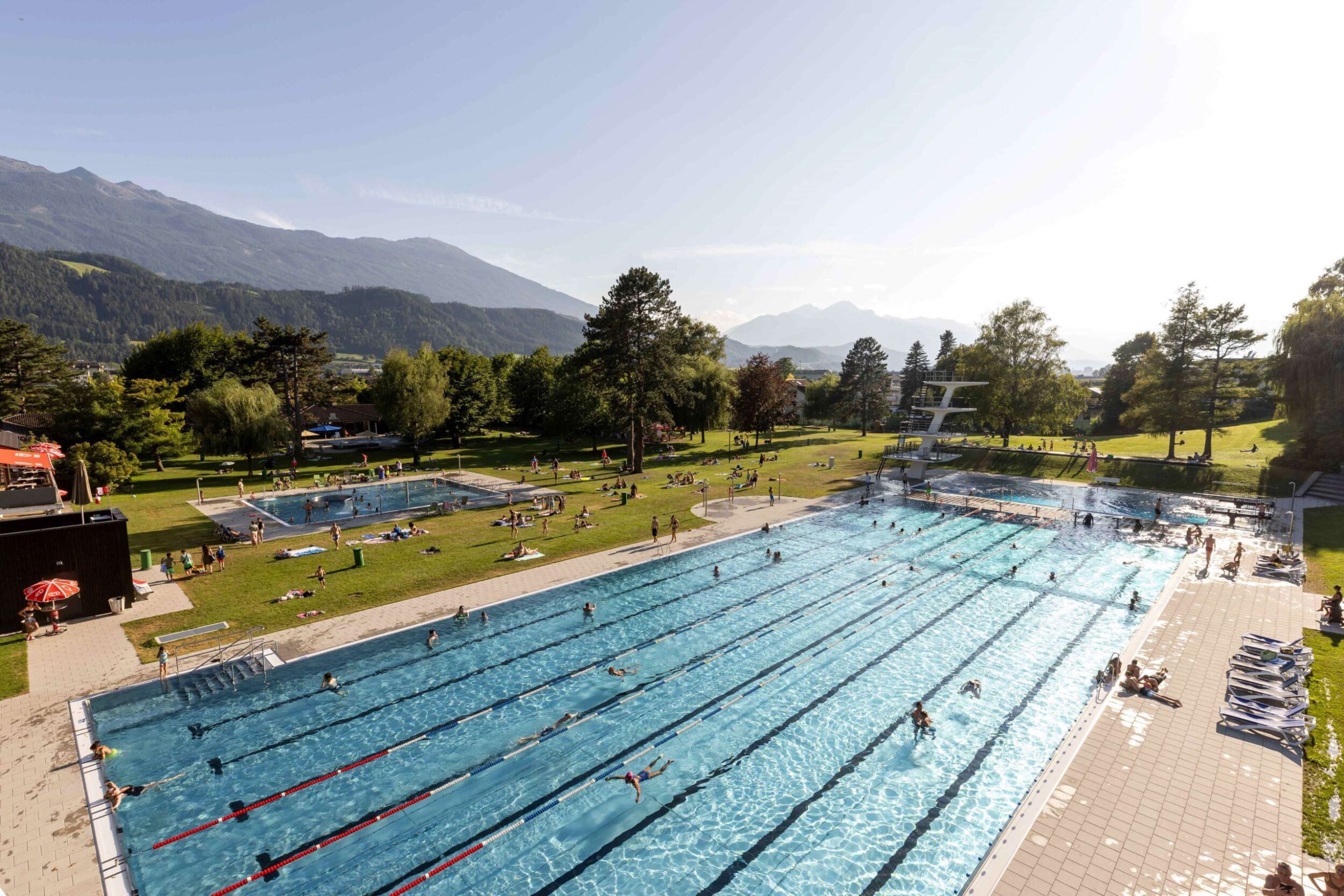 Außen-Schwimmbecken mit Schwimmbahnen, Sprungturm und Liegewiese vor Bergkulisse bei Sonnenschein