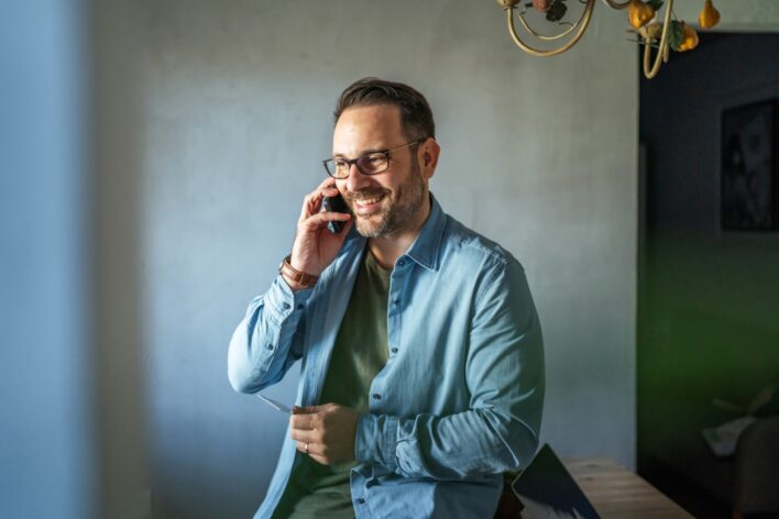 Mann mit Brille und blauem Hemd telefoniert lächelnd in hellem Raum.