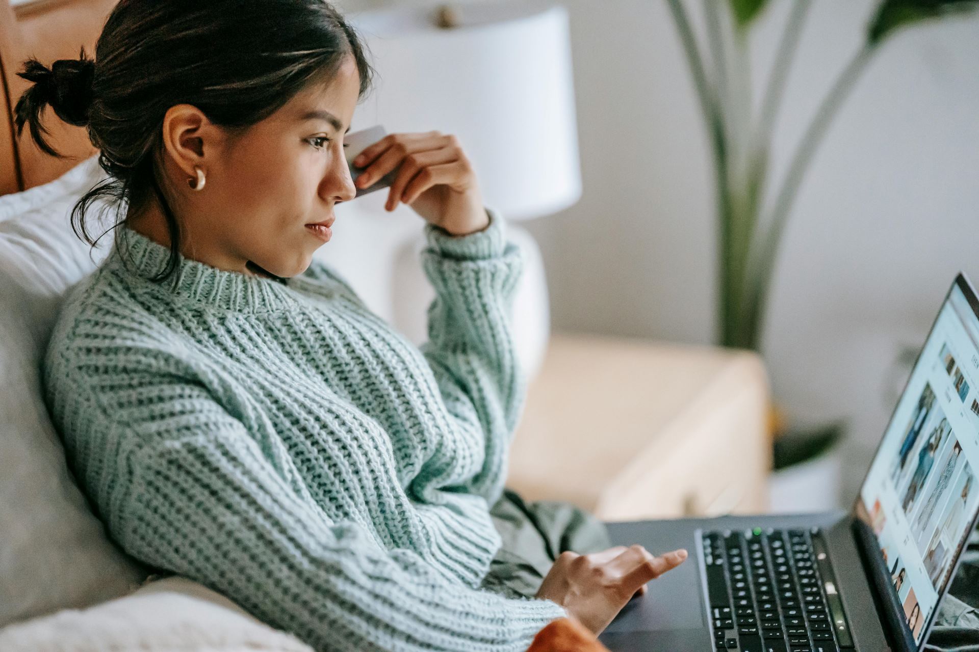 Frau mit Zopf und grauem Strickpullover sitzt auf Sofa und arbeitet konzentriert an Laptop.