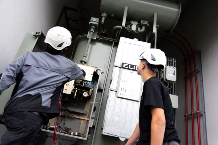 Zwei Personen mit Schutzhelmen stehen vor einem großen elektrischen Schaltgerät in einem Umspannwerk