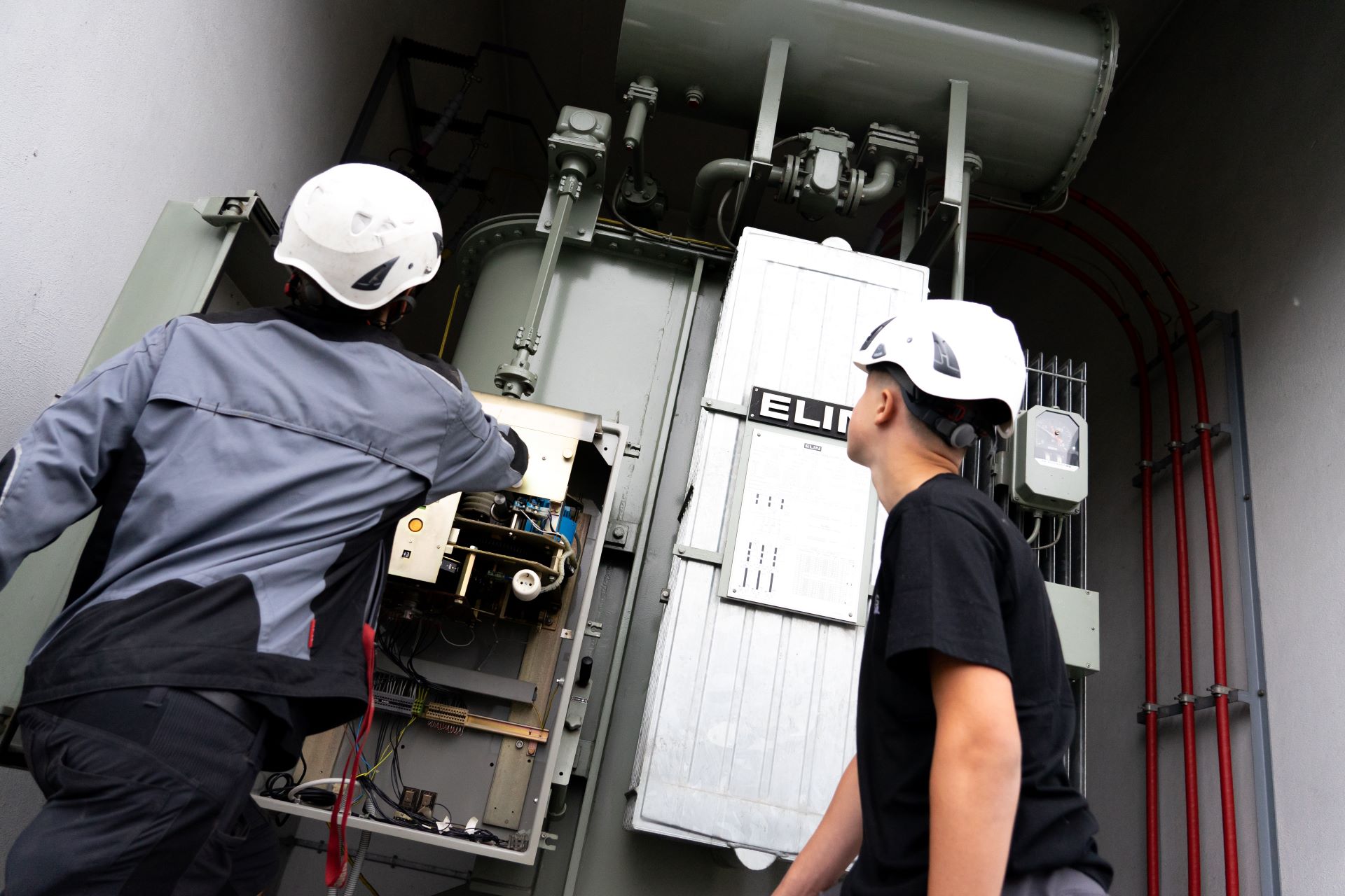 Zwei Personen mit Schutzhelmen stehen vor einem großen elektrischen Schaltgerät in einem Umspannwerk