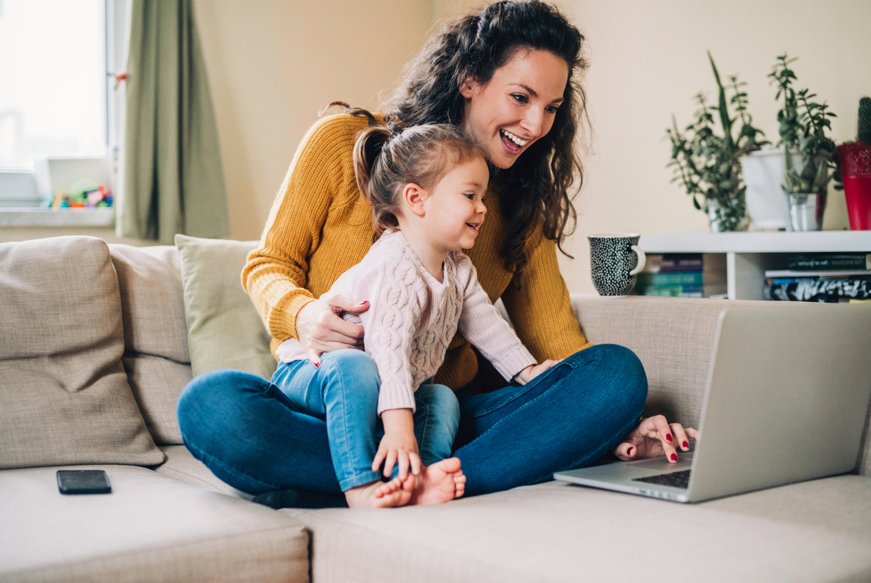 Eine Frau mit lockigen, braunen Haaren und gelbem Pullover sitzt auf einem hellen Sofa und hält ein kleines Kind auf dem Schoß. Beide schauen lächelnd auf einen geöffneten Laptop, der auf dem Sofatisch steht. Im Hintergrund stehen mehrere grüne Zimmerpflanzen auf einer Kommode, Tageslicht fällt durch ein Fenster mit grünen Vorhängen. Ein Smartphone liegt auf dem Sofa neben der Frau.