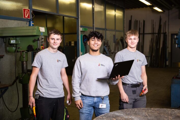 Drei junge Männer in grauen T-Shirts stehen in einer Werkstatt, einer hält einen Laptop, ein anderer einen Schraubenzieher