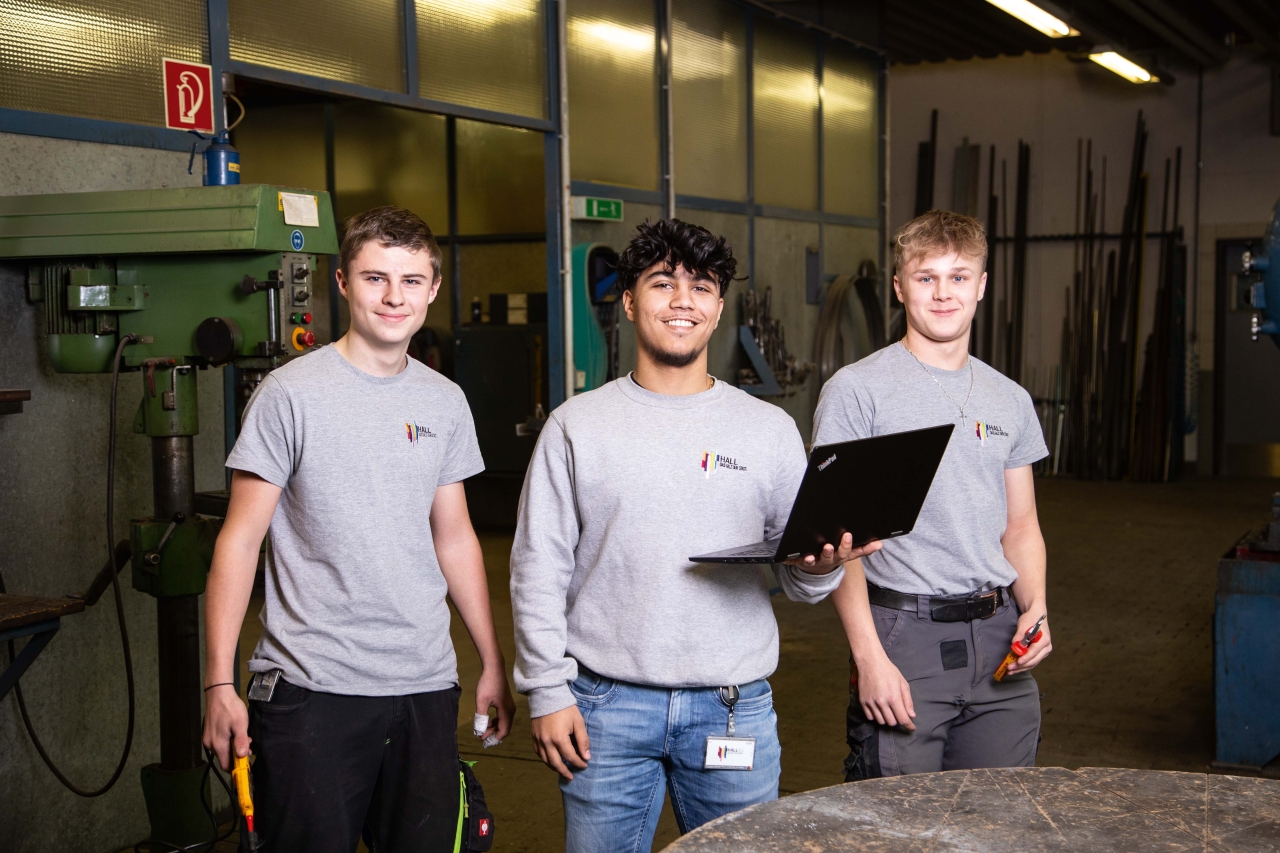 Drei junge Männer in grauen T-Shirts stehen in einer Werkstatt, einer hält einen Laptop, ein anderer einen Schraubenzieher