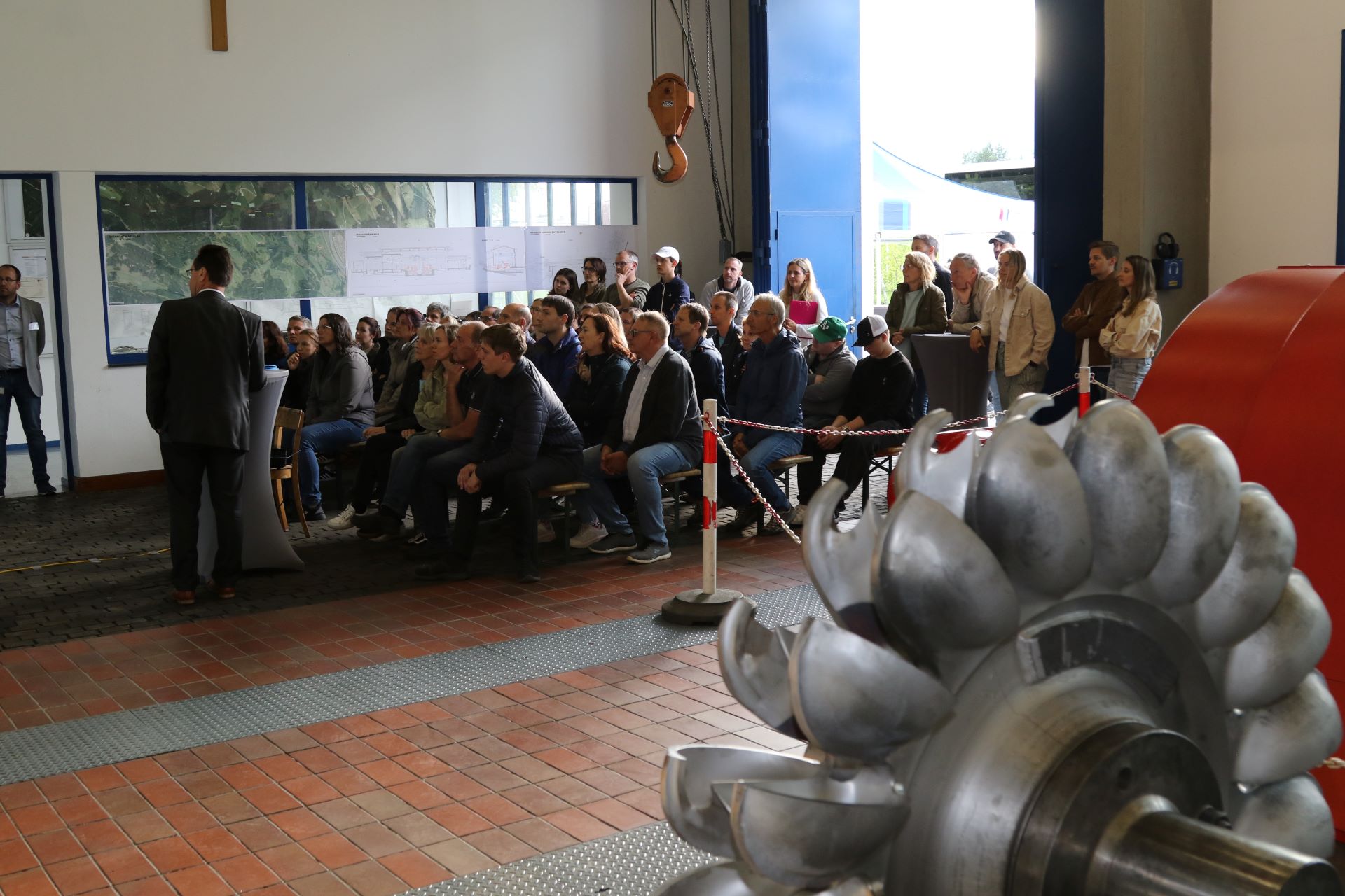 Gruppe von Menschen sitzt und steht in einer Halle vor einem Mann, der spricht, im Vordergrund ein großes Metallrad mit Schaufeln