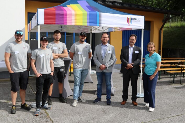 Gruppe von sieben Personen steht vor einem Pavillon mit buntem Dach und gelbem Gebäude im Hintergrund