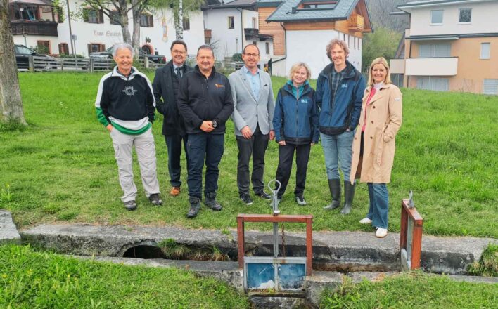 Sieben Personen stehen auf einer Wiese vor einem kleinen Wasserkanal mit einer Metallvorrichtung zur Wasserregulierung