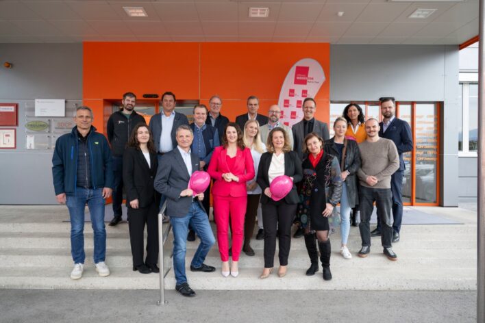 Gruppe von Menschen steht vor einem offenen Werkstor mit orangefarbener Fassade und hält pinkfarbene Helme