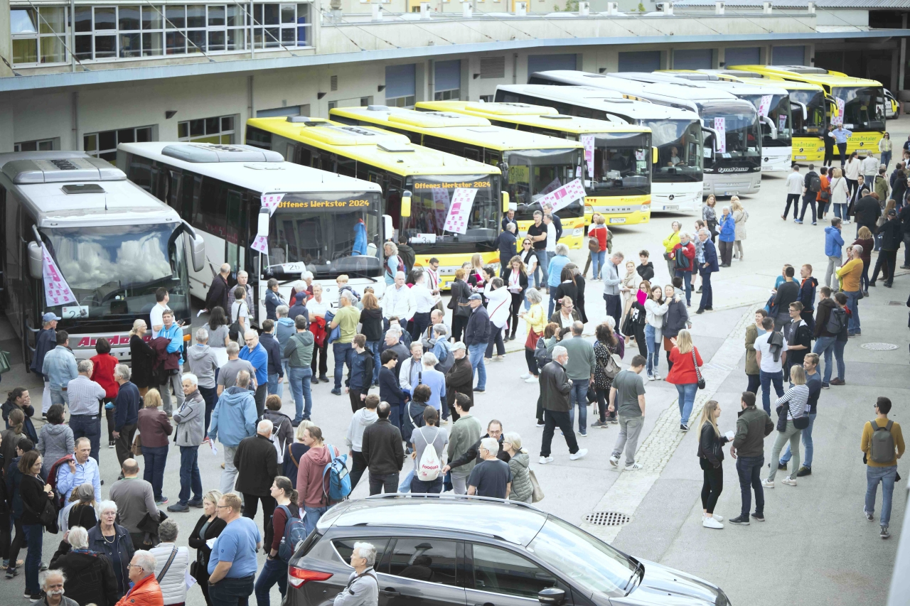 Menschenmenge vor einer Reihe geparkter Reisebusse an einem offenen Werkstor
