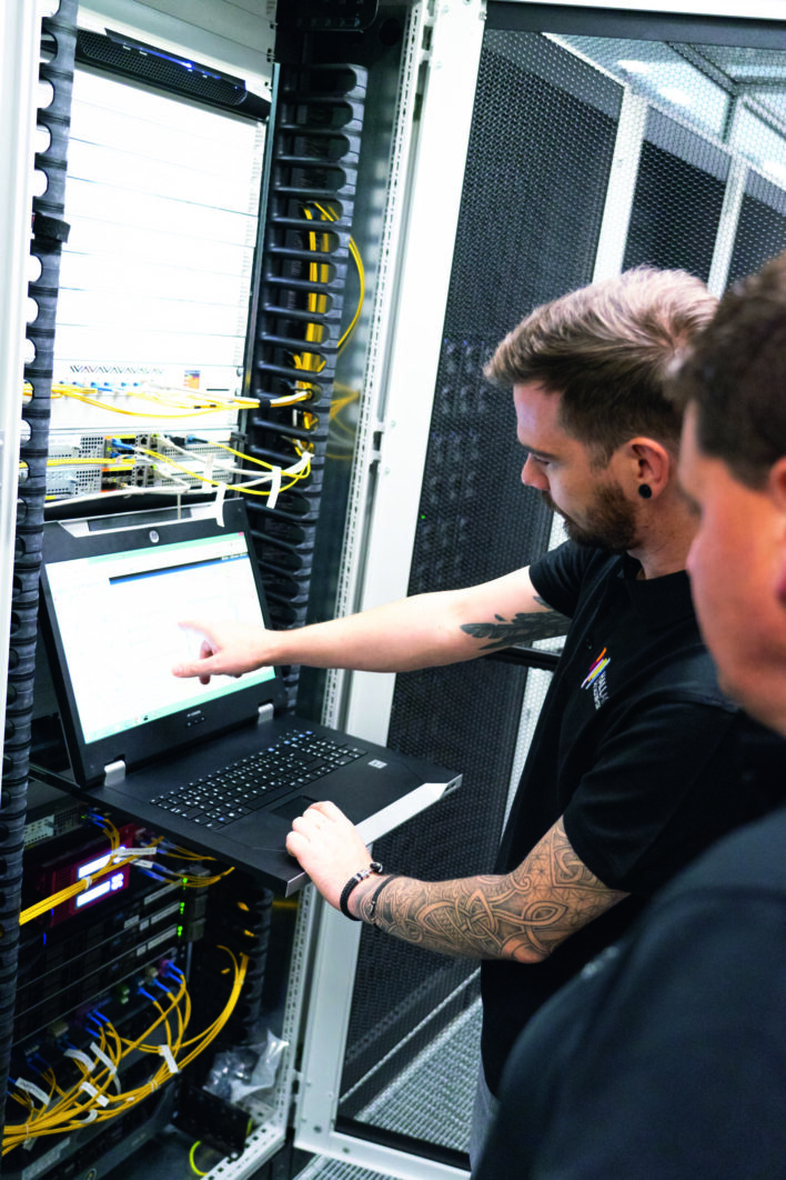 Zwei Männer stehen vor einem Serverrack, einer zeigt auf einen Bildschirm an einem Laptop im Rechenzentrum.