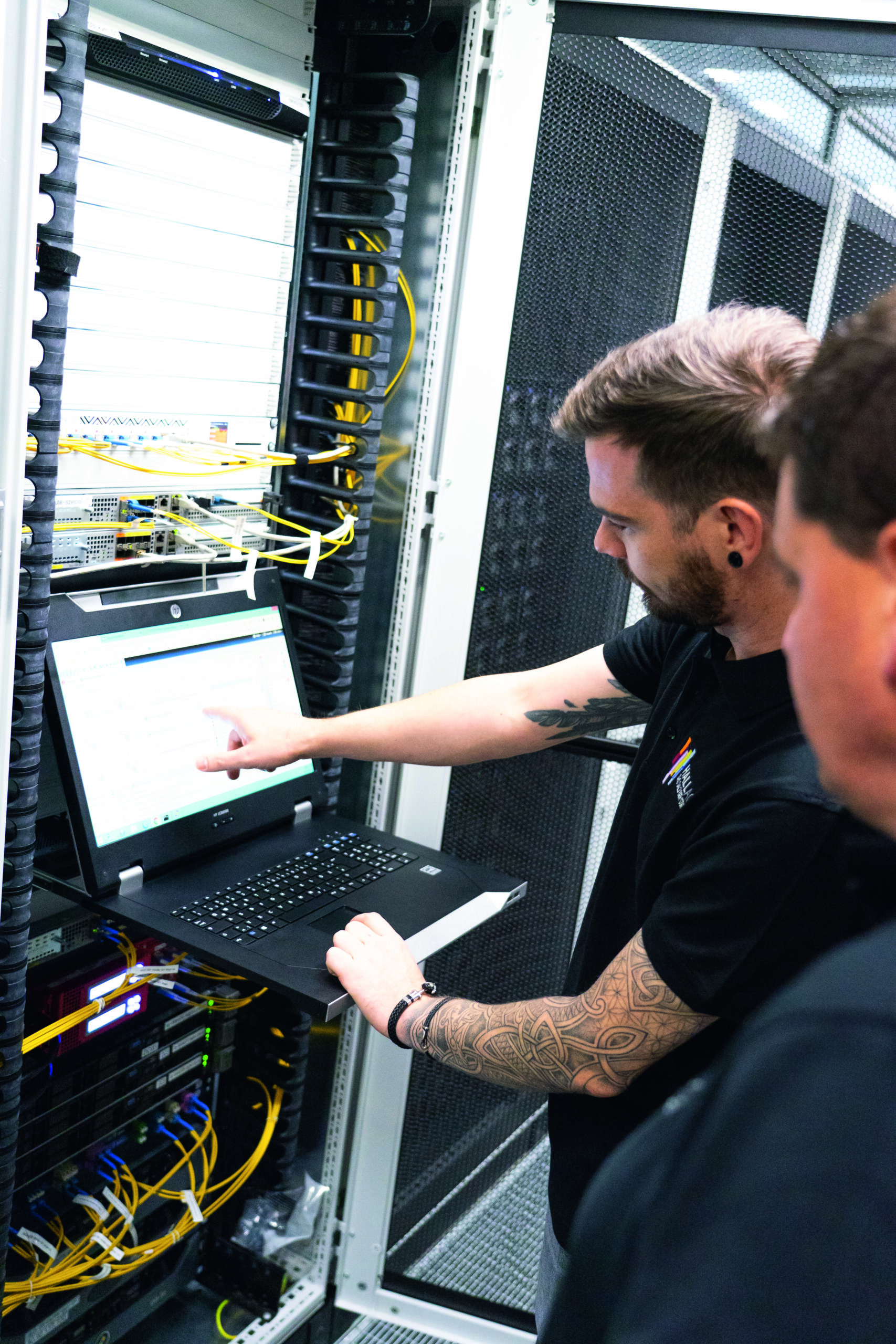 Zwei Männer stehen vor einem Serverrack, einer zeigt auf einen Bildschirm an einem Laptop im Rechenzentrum.