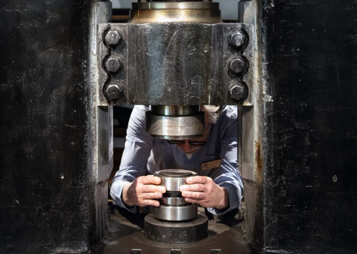 Person in weißem Hemd und Brille bedient eine große Metallpresse und hält ein zylindrisches Metallteil