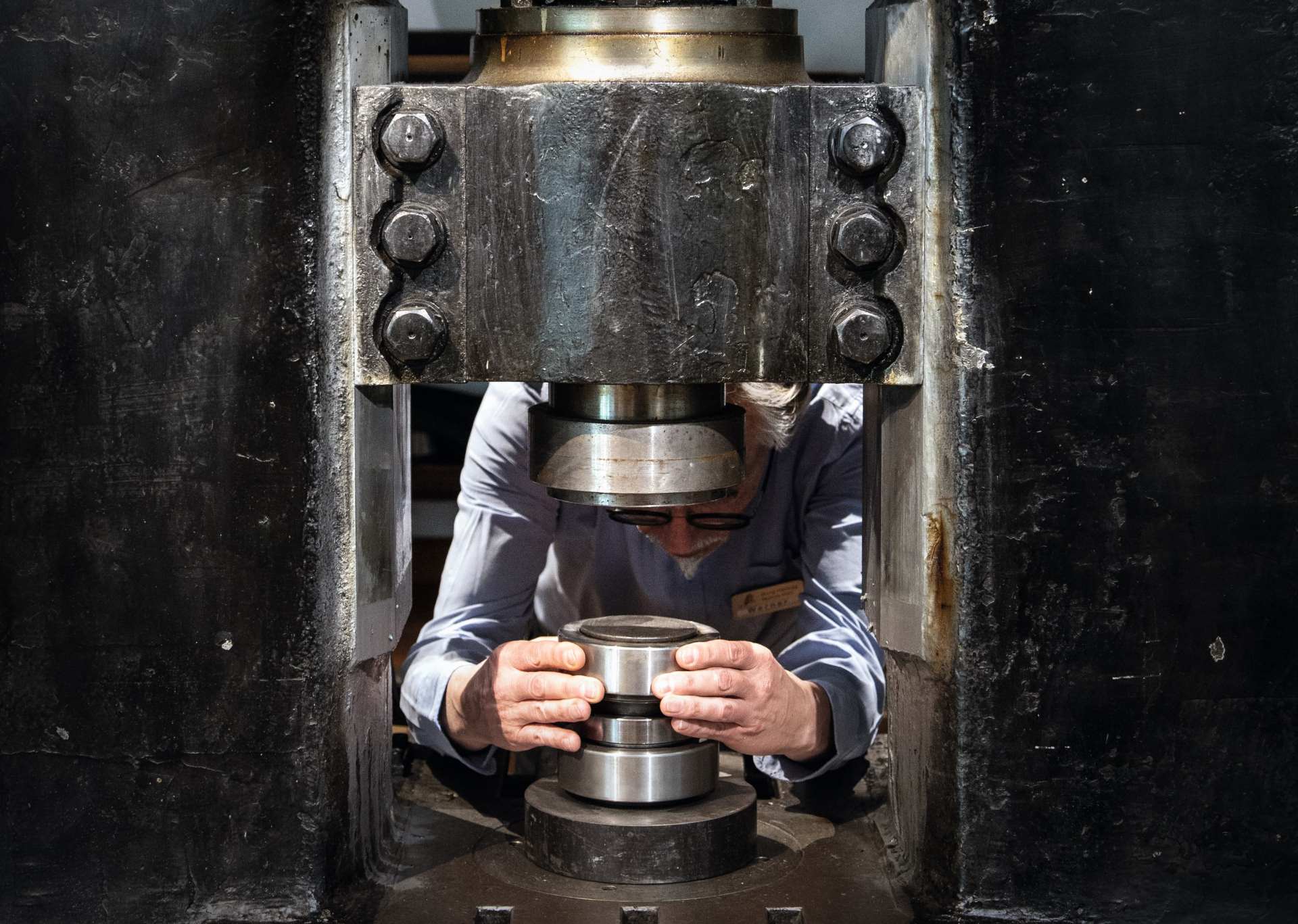 Person in weißem Hemd und Brille bedient eine große Metallpresse und hält ein zylindrisches Metallteil