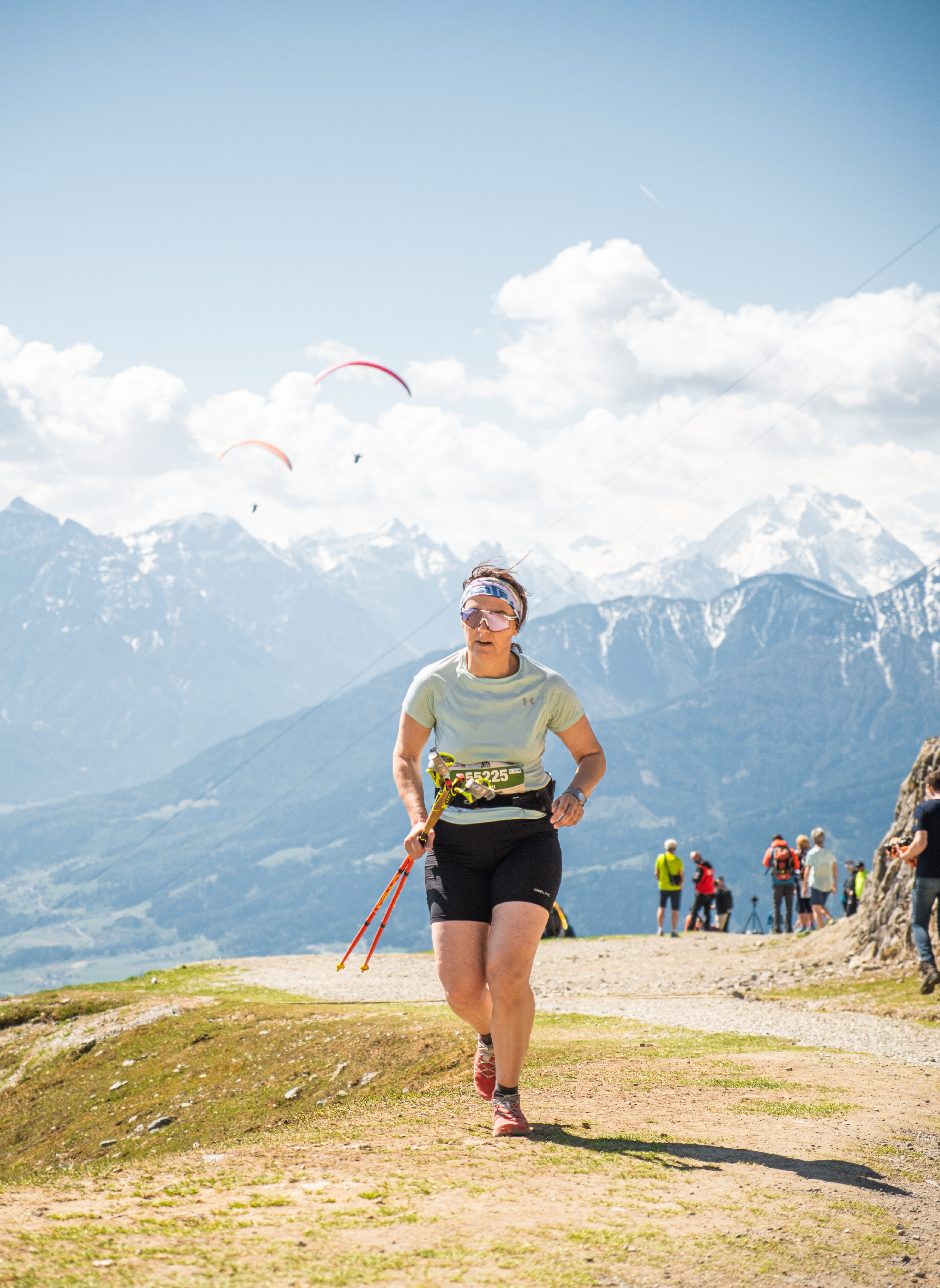 Eine Trailrunnerin läuft mit Stöcken auf einem alpinen Pfad an der Nordkette über Innsbruck, während Gleitschirmflieger im Hintergrund vor der Bergkulisse kreisen.