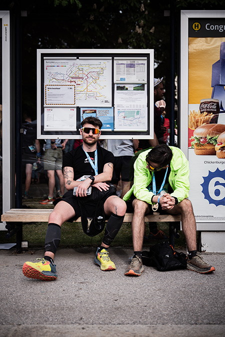 Zwei erschöpfte Trailrunner mit Finisher-Medaillen ruhen sich nach dem Innsbruck Alpine Trailrun Festival an einer Bushaltestelle in Innsbruck aus.