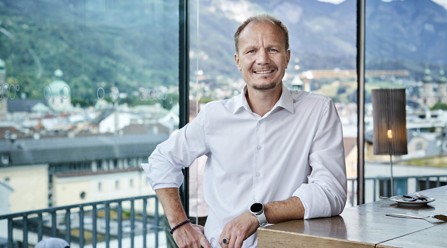 Porträt von Johannes Anzengruber, Bürgermeister von Innsbruck, in einem weißen Hemd vor einer Fensterfront mit Blick auf die Innsbrucker Altstadt und die Nordkette.