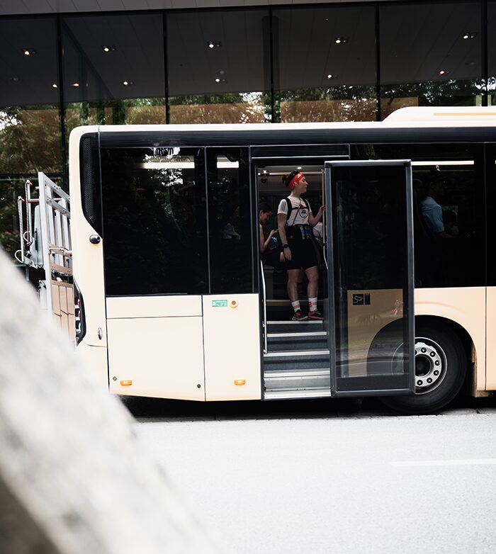 A beige bus with open doors is parked by the road. Two people wearing backpacks and hats are standing inside the bus near the entrance. The background shows reflections on a glass building.