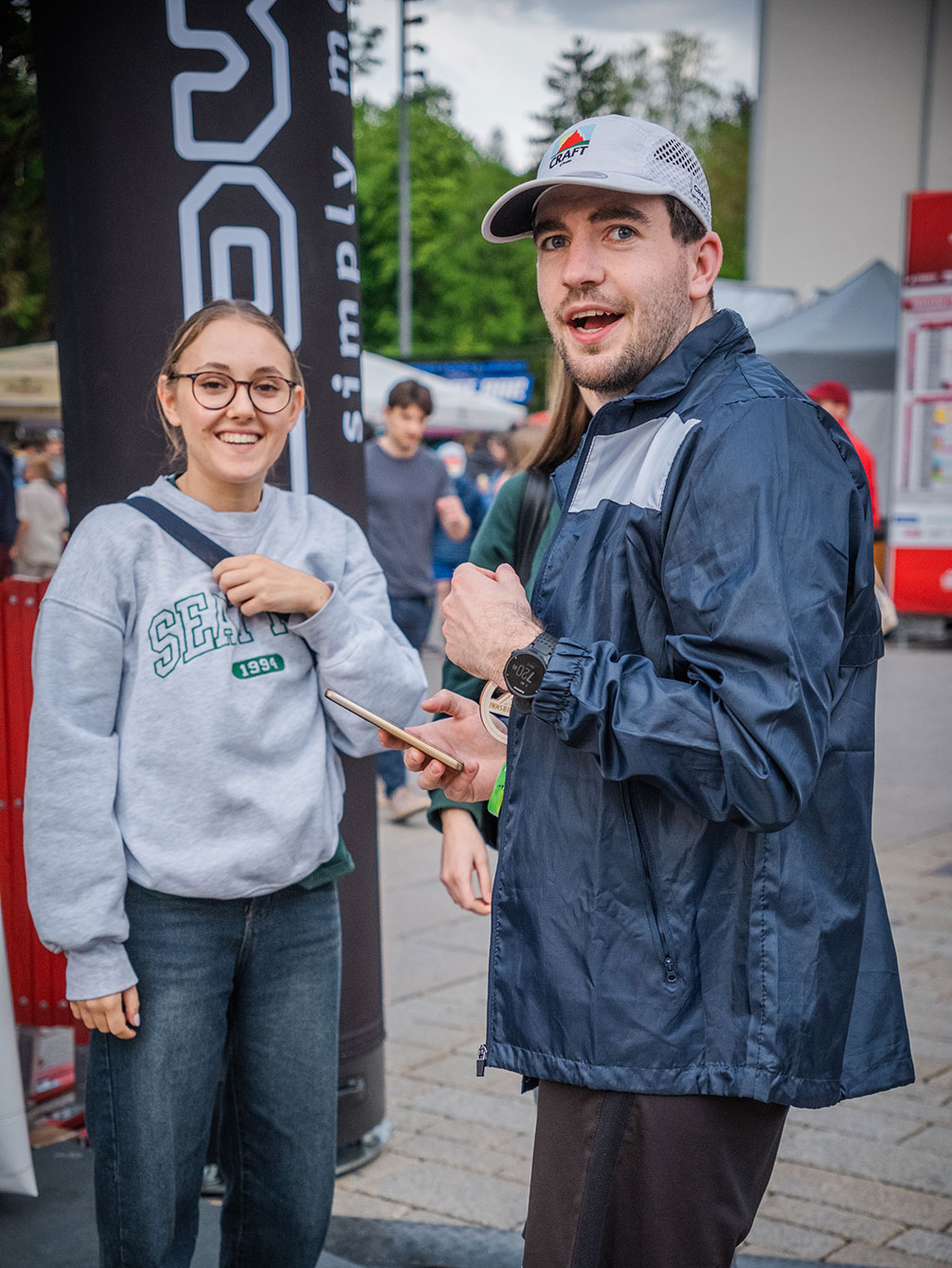 Eine junge Frau im grauen Sweatshirt und ein junger Mann mit Kappe lächeln in die Kamera in der Expo-Area des IATF in Innsbruck.