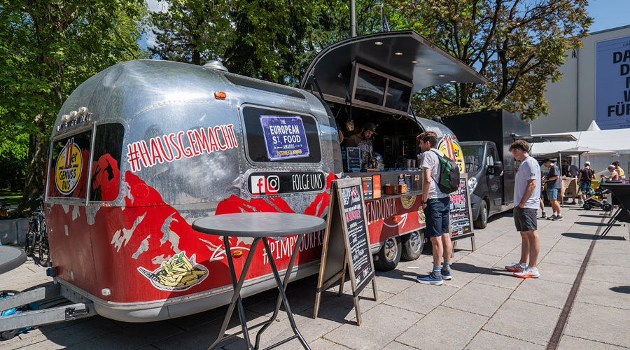 Ein silberner Foodtruck mit der Aufschrift ‚Hausgemacht‘ steht auf dem Eventgelände in Innsbruck, davor warten Besucher in der Sonne.