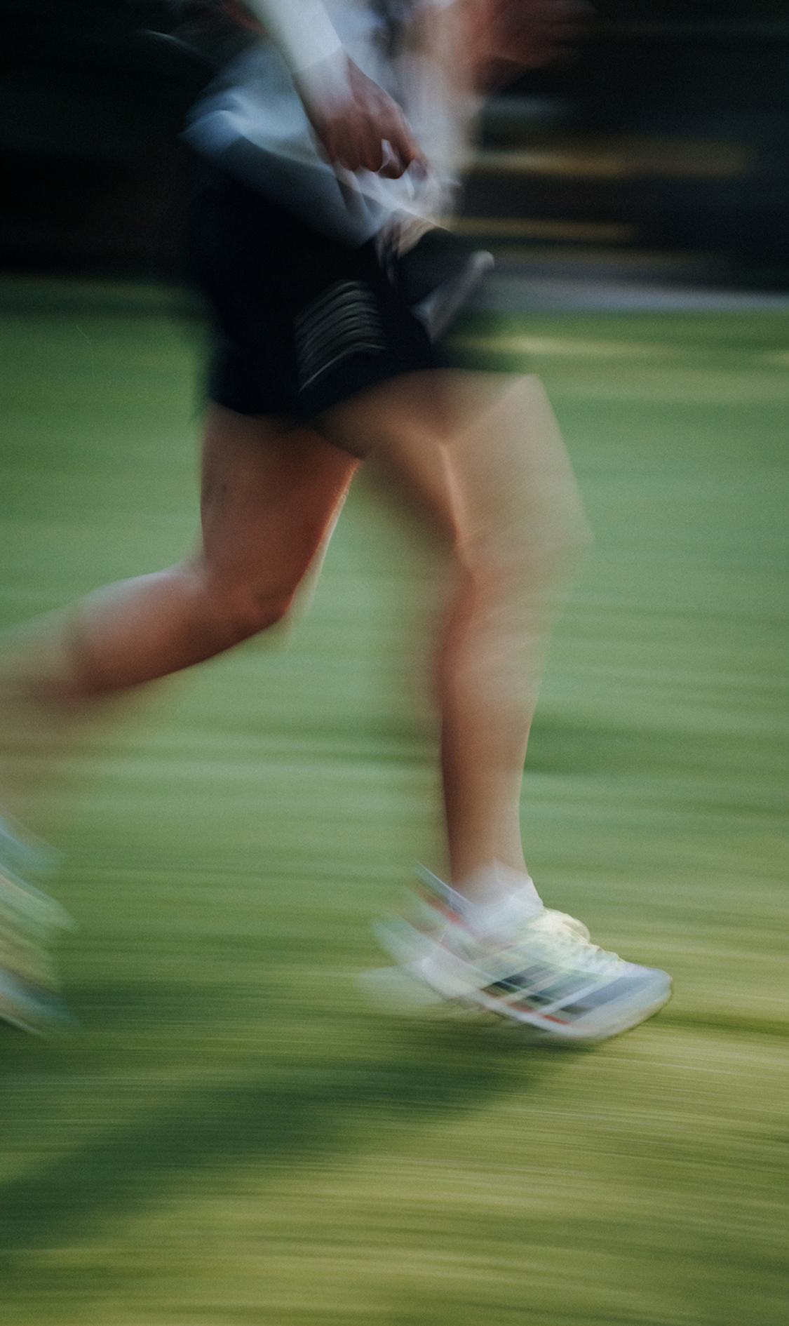 Bewegungsunschärfe zeigt die Beine einer laufenden Person in Sportkleidung auf einer grünen Wiese; weiße Laufschuhe berühren den Boden im schnellen Schritt.