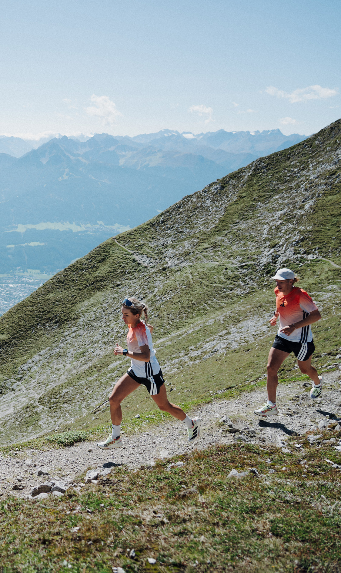 Zwei Läuferinnen oder Läufer laufen auf einem schmalen Bergpfad entlang eines steilen Hangs; tief unten im Tal ist eine Stadt mit Flusslauf zu sehen.