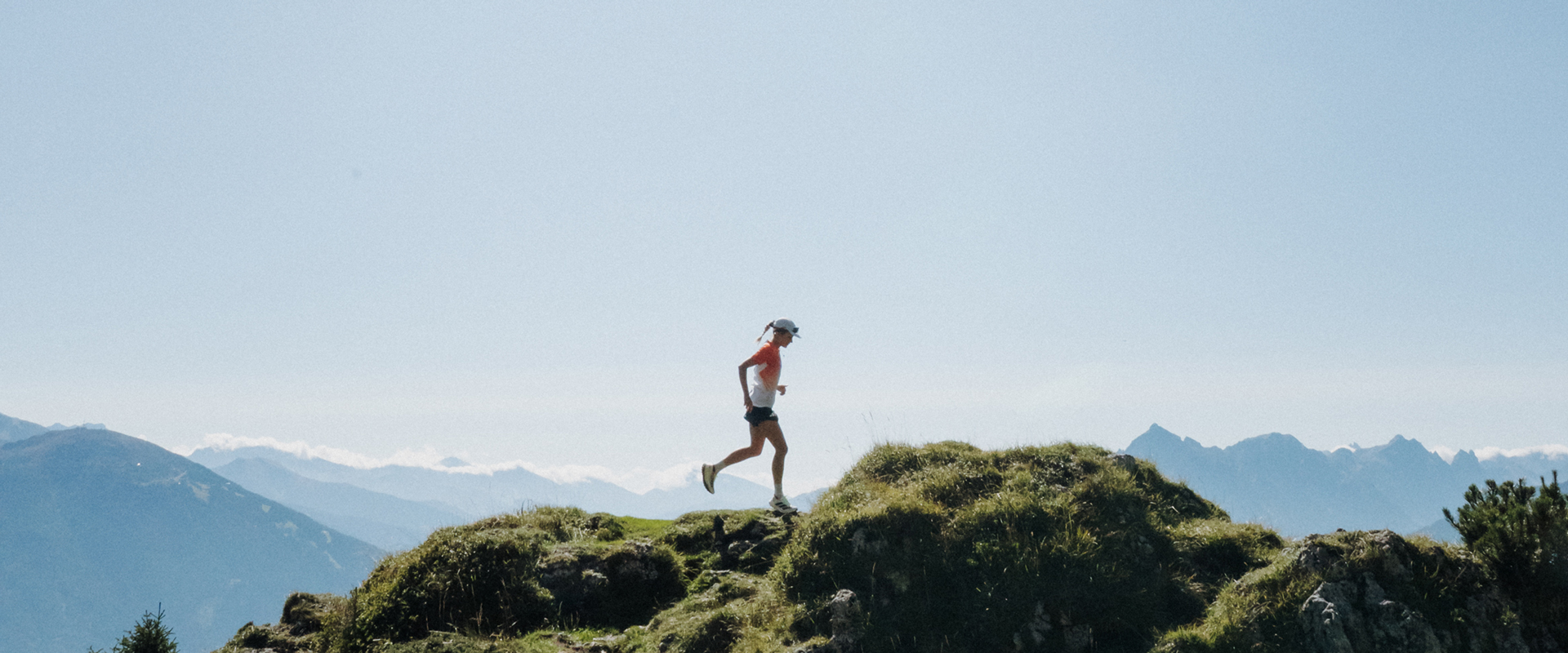 Eine Läuferin läuft über einen schmalen Bergkamm mit grasbewachsenen Hügeln; im Hintergrund sind weite Bergketten unter einem klaren Himmel zu sehen.