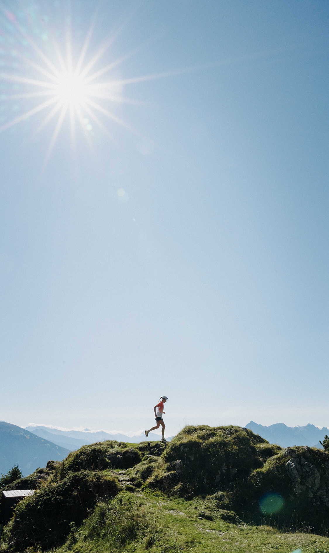 Eine Läuferin läuft über einen schmalen Bergkamm mit grasbewachsenen Hügeln; im Hintergrund sind weite Bergketten unter einem klaren Himmel zu sehen.