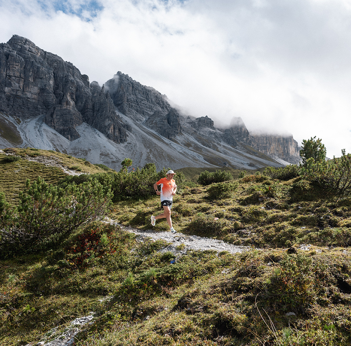 Ein Läufer läuft auf einem schmalen Bergpfad durch eine alpine Landschaft; im Hintergrund erheben sich felsige Gipfel, teils von Wolken umhüllt.