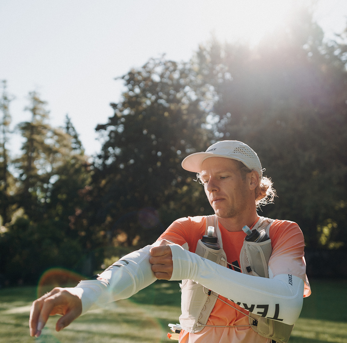 Ein Läufer in Sportkleidung zieht sich in einer sonnigen, grünen Umgebung einen Armstulpen über; er trägt eine Laufweste mit Trinkflaschen.