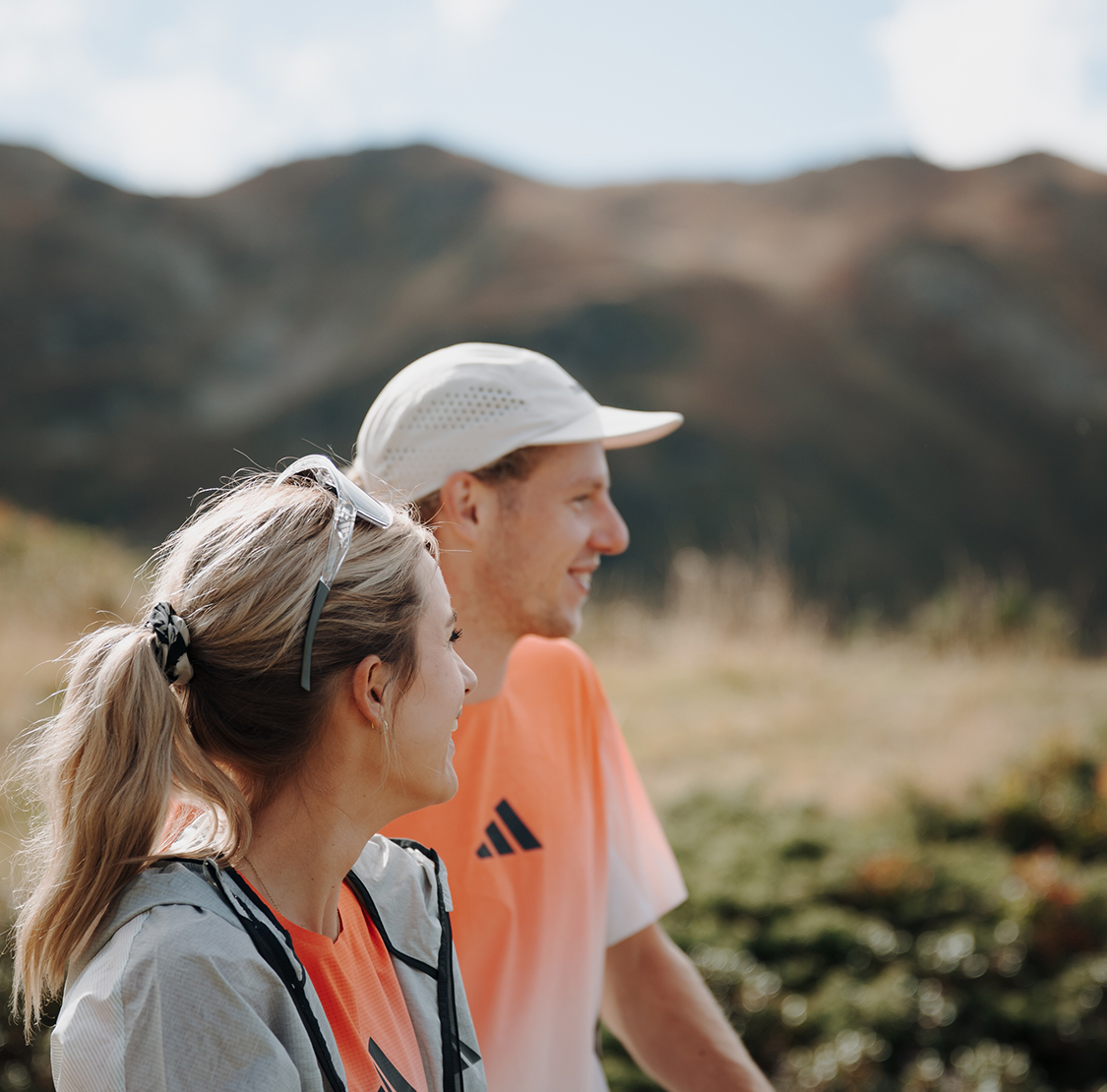 Zwei sportlich gekleidete Personen stehen in einer bergigen Landschaft und blicken lächelnd in die Ferne; im Hintergrund sind Hügel und eine weite Naturkulisse zu sehen.