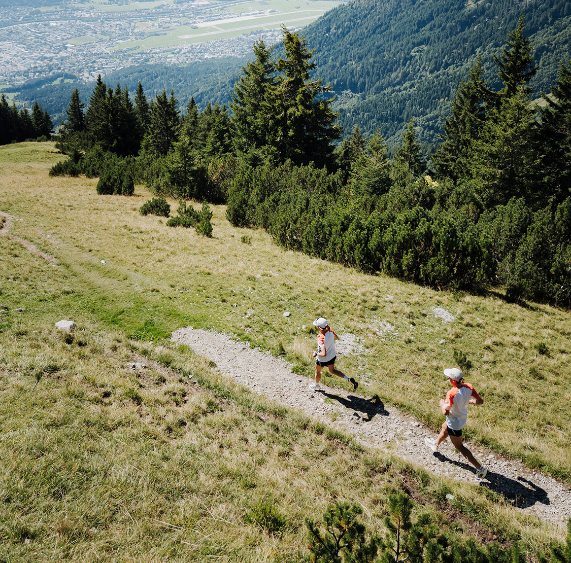 Zwei Läufer laufen auf einem schmalen Bergpfad durch eine grüne Hügellandschaft; im Hintergrund sind Wälder und ein Tal mit Stadtansicht zu sehen.
