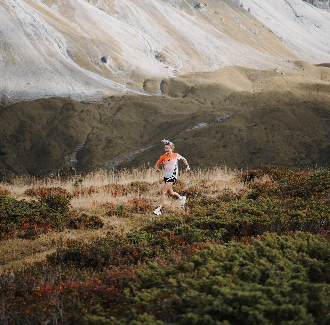 Eine Läuferin läuft über einen schmalen Pfad durch eine alpine Landschaft mit Gras und Sträuchern; im Hintergrund erheben sich karge Berghänge.