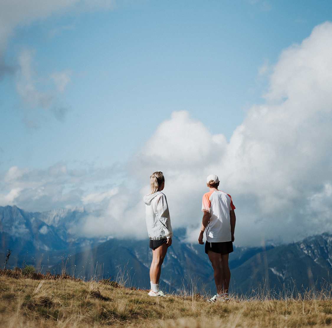 Zwei sportlich gekleidete Personen stehen auf einem Bergkamm und blicken auf eine weite Berglandschaft mit Wolken und Gipfeln im Hintergrund.
