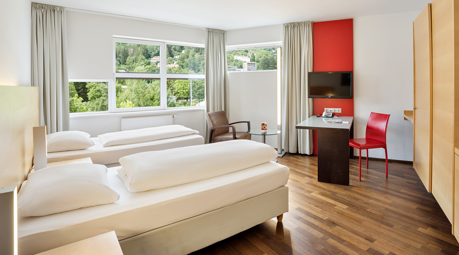 Ein modernes, helles Hotelzimmer im Austria Trend Hotel in Innsbruck mit zwei Einzelbetten, Parkettboden, einem roten Designerstuhl und großem Fenster mit Blick auf bewaldete Hänge.