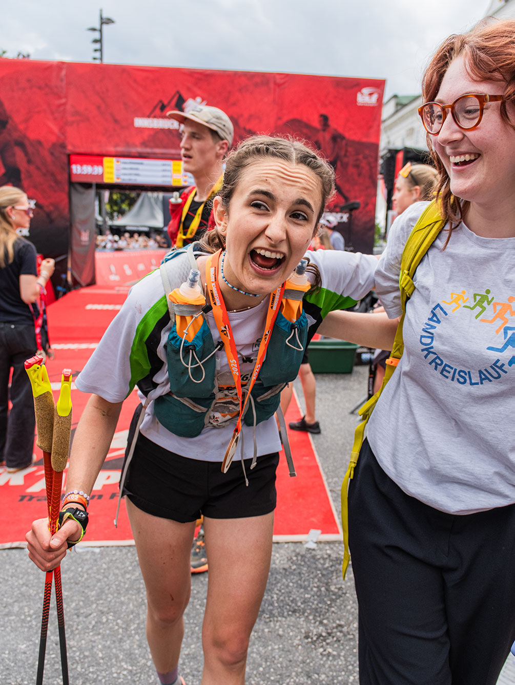 Zielbereich des Innsbruck Alpine Trailrun Festivals: Eine Läuferin mit Medaille, Laufweste und Trailrunning-Stöcken feiert ihren Erfolg vor dem roten IATF-Zielbogen. Im Hintergrund ist die Zeitanzeige und das Event-Branding zu sehen.