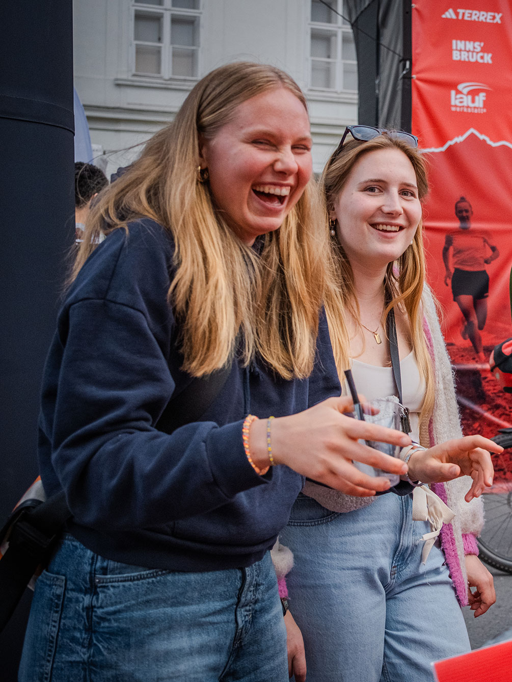Zwei lachende junge Frauen genießen die Atmosphäre in der Expo-Area des IATF in Innsbruck vor dem roten Zielbogen.