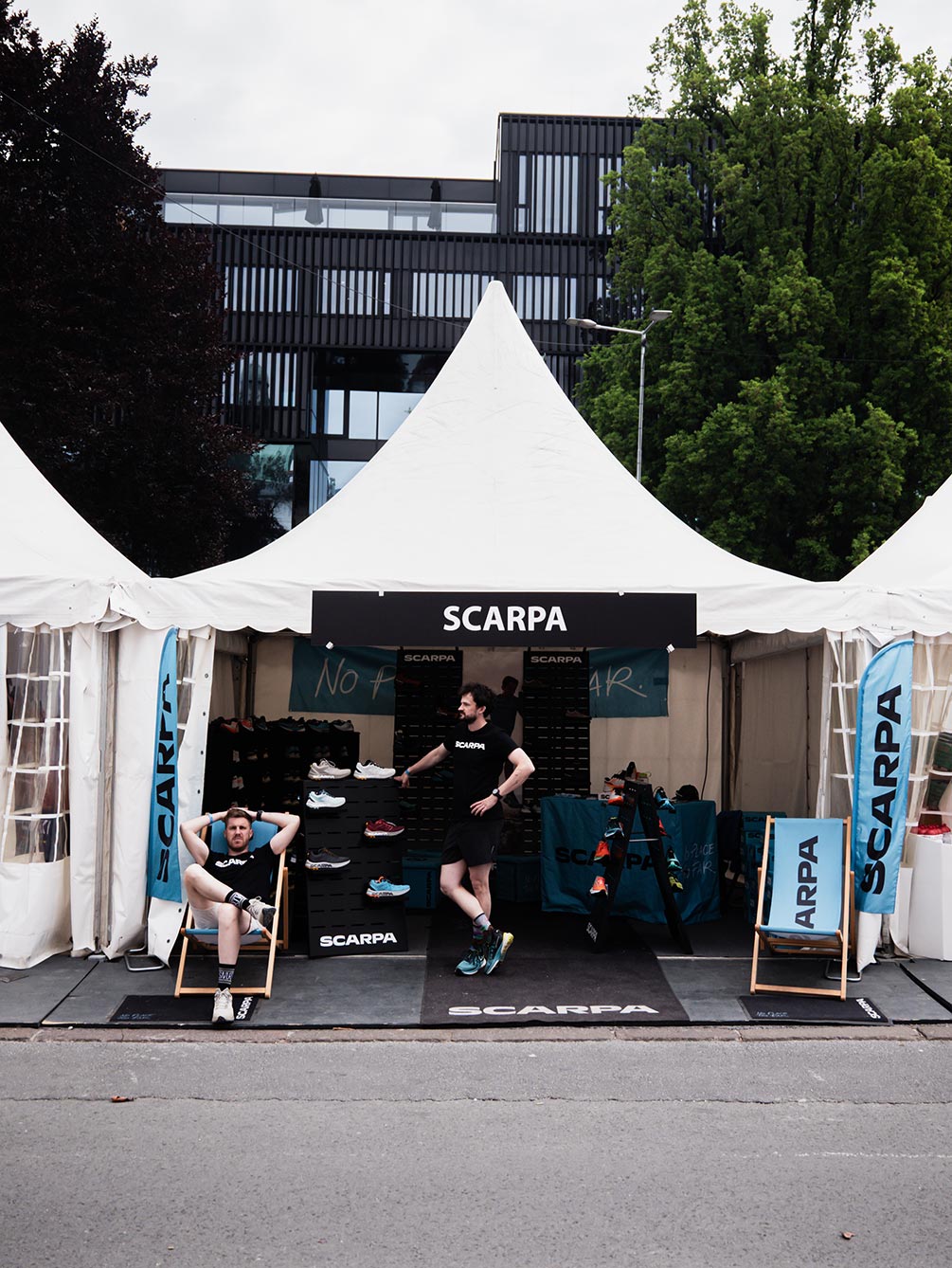 Ein weißes Ausstellerzelt der Marke SCARPA auf der Outdoor-Expo des Innsbruck Alpine Trailrun Festivals mit präsentierten Trailrunning-Schuhen und entspannten Mitarbeitern in Liegestühlen.