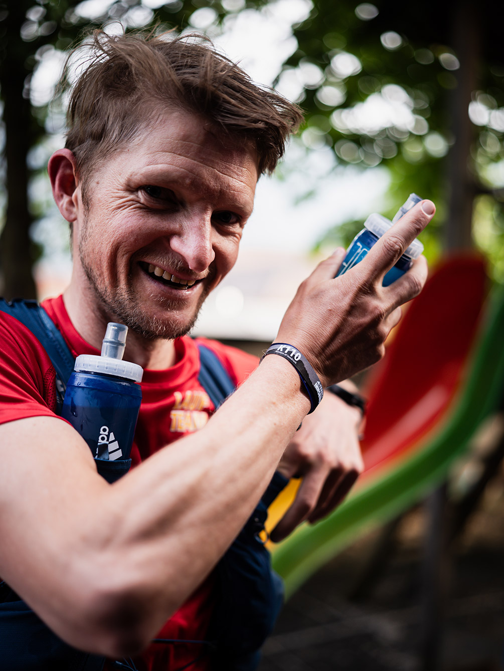 Porträt eines lächelnden Trailrunners mit K110-Armband, der Softflasks von Adidas in der Hand und an der Laufweste hält.