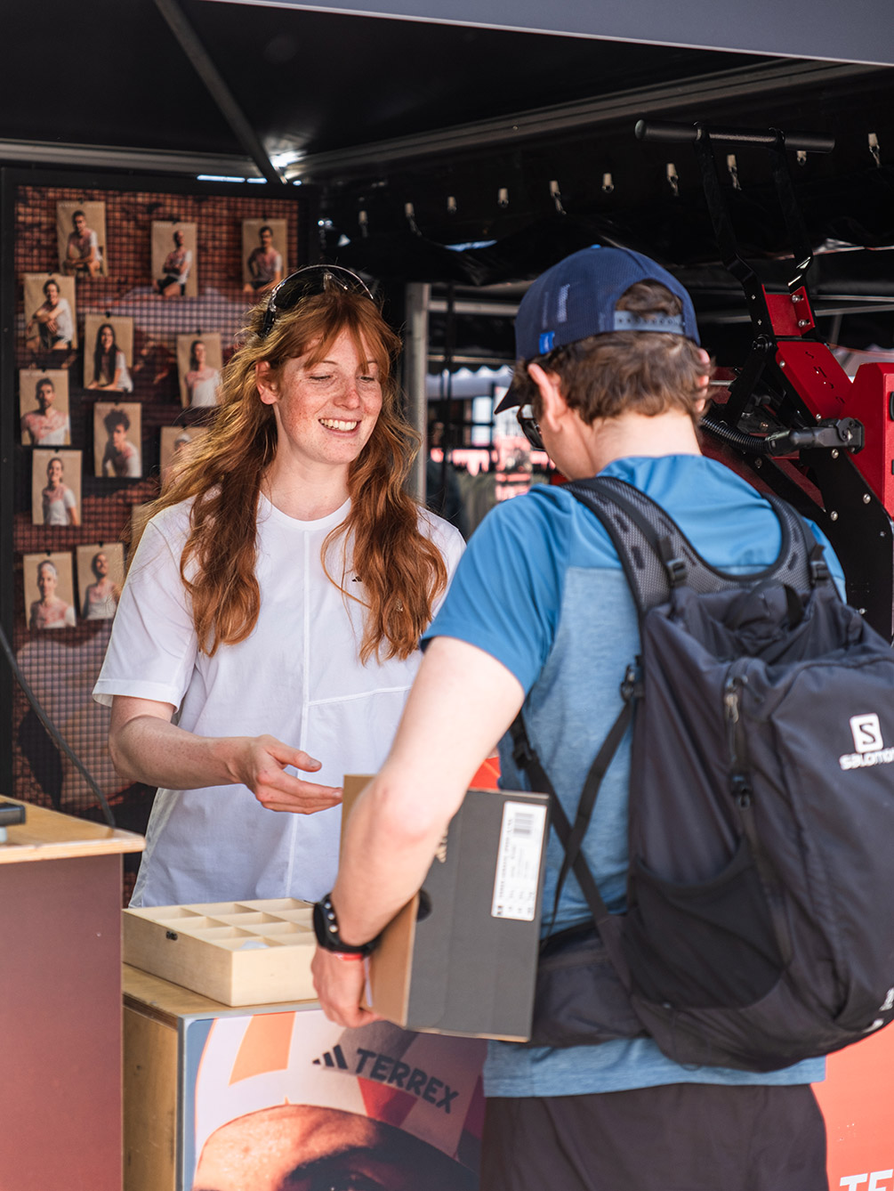 Eine freundliche Mitarbeiterin am Adidas Terrex Stand der IATF Expo übergibt einem Teilnehmer ein Produktpaket; im Hintergrund ist eine Wand mit Athleten-Porträts zu sehen.