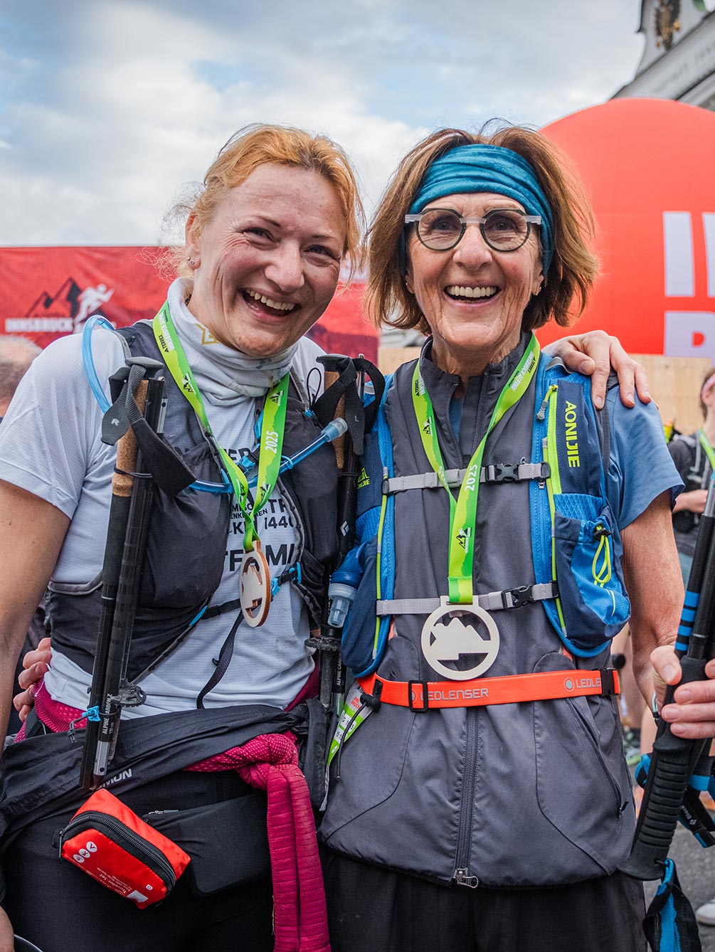 Zwei strahlende Läuferinnen mit Finisher-Medaillen und Trailrunning-Ausrüstung umarmen sich nach dem Zieleinlauf beim Innsbruck Alpine Trailrun Festival.