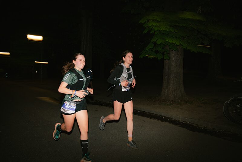 Zwei junge Frauen beim nächtlichen Lauf während des K7 Nighttrails des Innsbruck Alpine Trailrun Festivals auf einem von Bäumen gesäumten Weg.