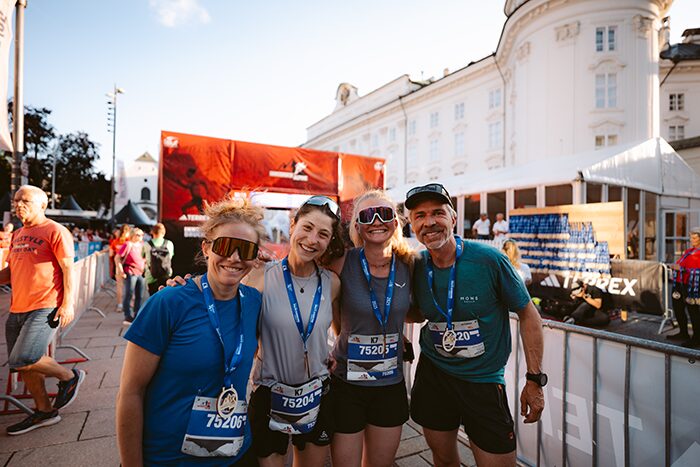 Eine Gruppe von vier lachenden Trailrunnern mit Medaillen um den Hals posiert nach dem Rennen vor dem weißen Gebäude der Hofburg in Innsbruck.“