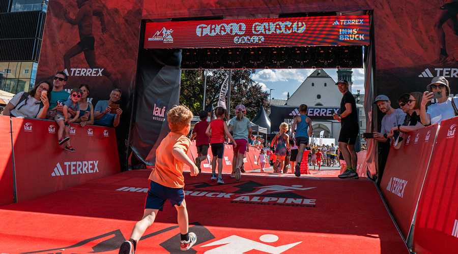 Eine Gruppe von Kindern läuft beim Innsbruck Alpine Trailrun Festival unter einem großen digitalen Torbogen mit der Aufschrift ‚Trail Champ‘ hindurch.
