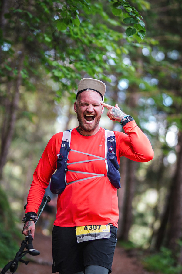 Ein bärtiger Trailrunner in einem leuchtend orangefarbenen Langarmshirt und blauer Laufweste lacht laut und zeigt sich beim Innsbruck Alpine Trailrun Festival im Wald enthusiastisch an den Kopf.