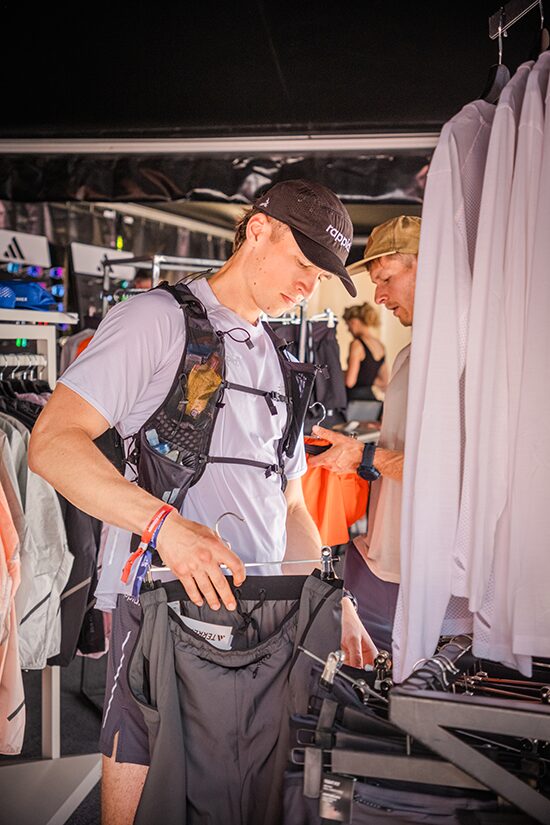 A man wearing a cap and running vest browses through clothing on a rack in a sports apparel store, while another person looks at clothing in the background.