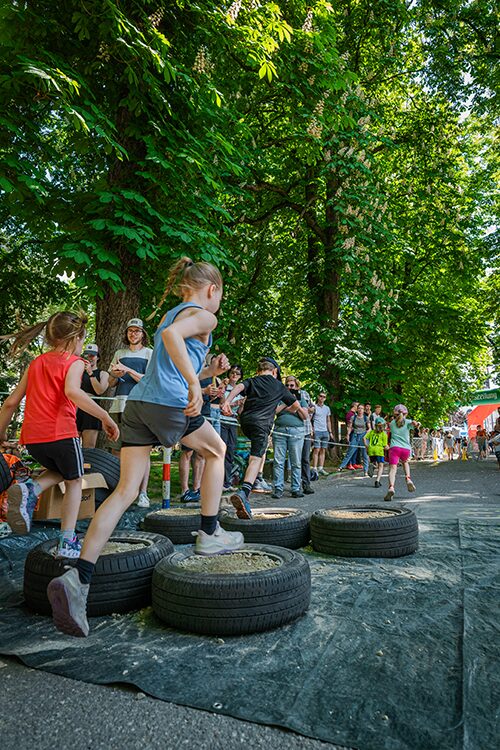 Children run and jump over tires arranged on the ground as part of an outdoor obstacle course, with a crowd of spectators watching under tall, leafy trees.