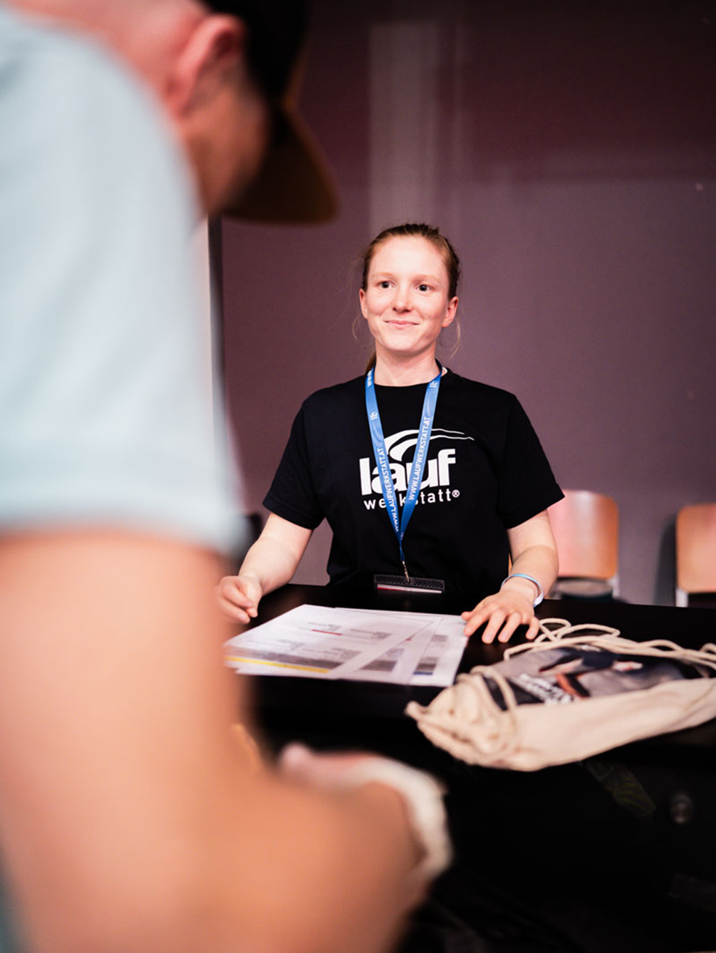 Eine lächelnde Volunteer-Mitarbeiterin der Laufwerkstatt am Counter für die Startnummernausgabe beim IATF in Innsbruck während der Unterlagenprüfung.