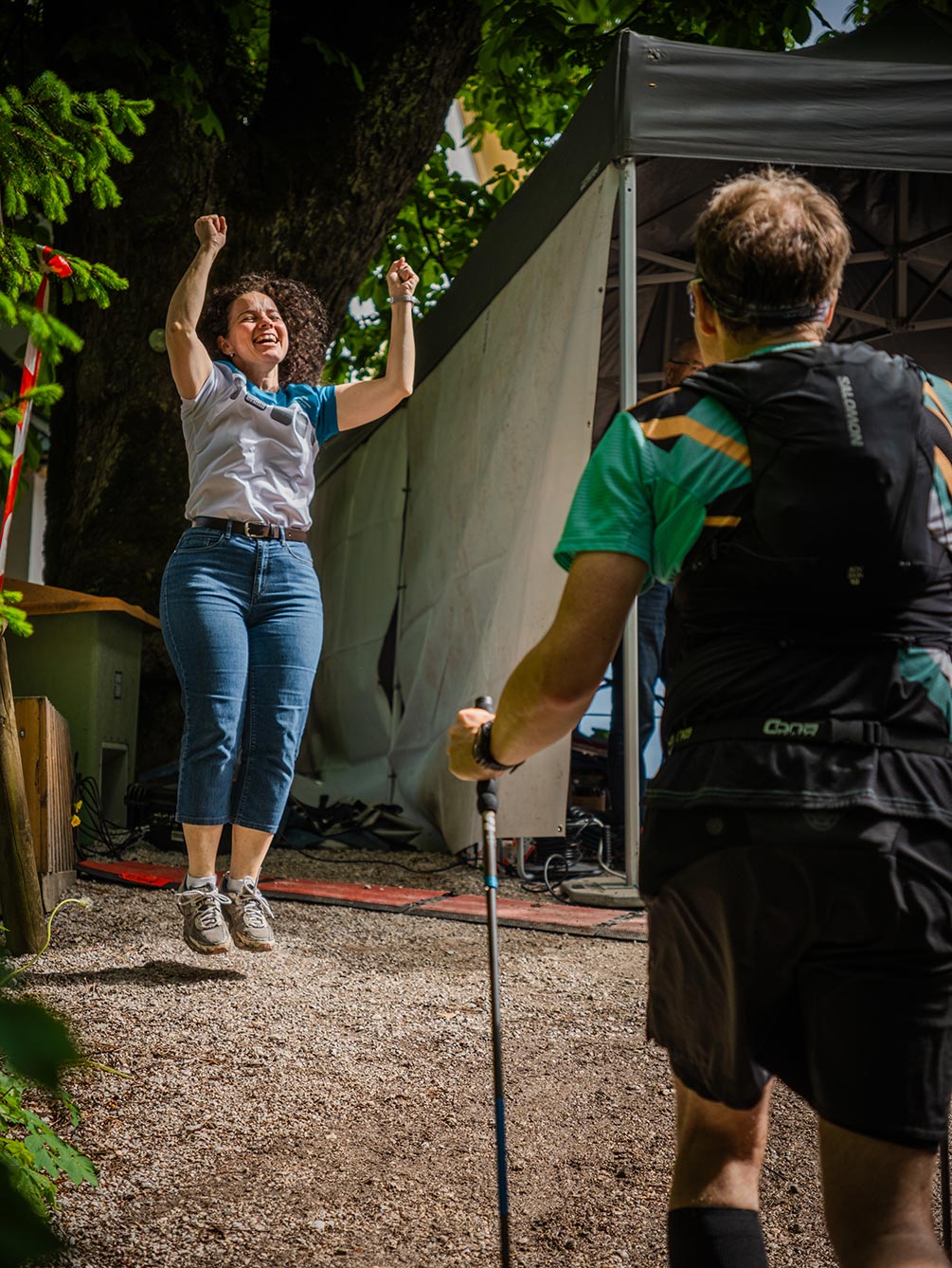 Eine jubelnde Zuschauerin springt vor Freude in die Luft, während ein Trailrunner mit Stöcken an einem Verpflegungspunkt des IATF vorbeiläuft.